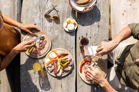 Drei Personen essen an einem Holztisch draußen verschiedene regionale Speisen von Tellern und schneiden Brot und Wurst.