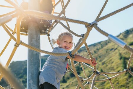 Ein lachendes Kind klettert an einem Seilnetz im Freien vor einer sonnigen Bergkulisse.