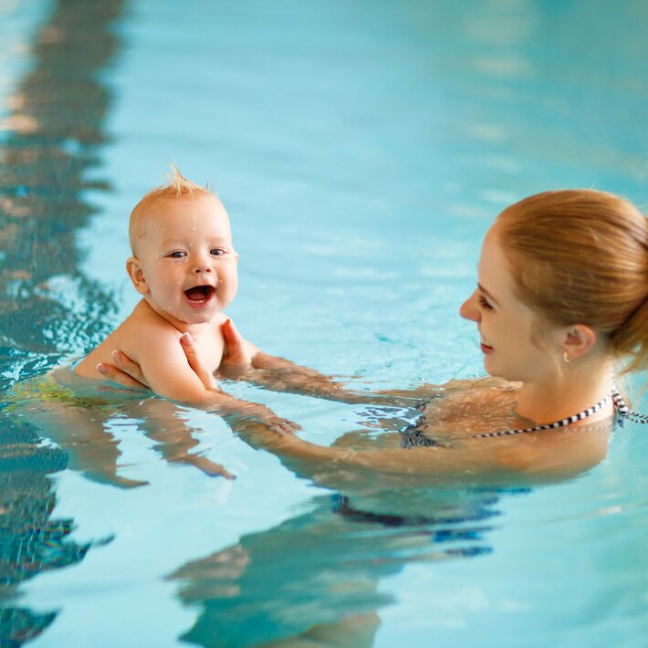 A mother holds her baby in the water of an indoor pool and smiles at the child.