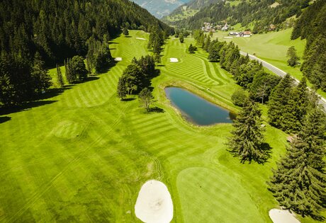 Ein gepflegter Golfplatz mit Wasserhindernis, Bunkern und fairwayartigen Bahnen liegt in einer grünen Berglandschaft.