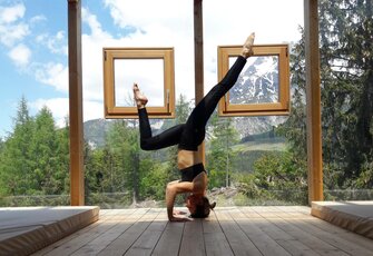 A woman is doing a forearm handstand on a wooden terrace in front of large windows overlooking the mountains.