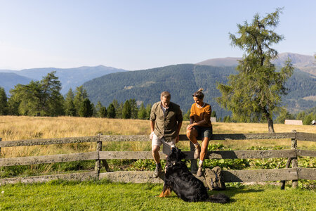 Zwei Wanderer sitzen an einem Holzzaun mit Hund vor einer Bergkulisse in sonniger Sommerlandschaft.