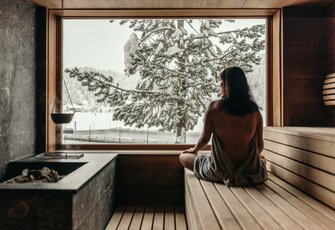 A woman sits in the sauna and looks through the large window toward the snowy lake and mountains.