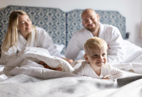 A child lies smiling on the bed while the parents sit in the background in the hotel room.