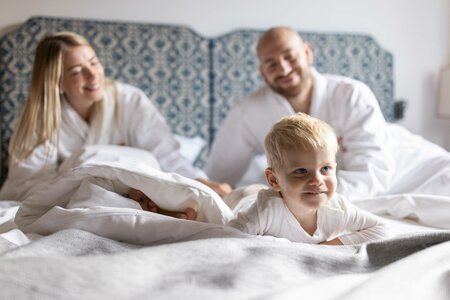 Ein Kind liegt lächelnd im Bett, während die Eltern im Hintergrund im Hotelzimmer sitzen.