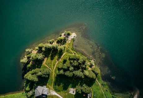 Blick von oben auf eine bewaldete Landzunge mit Wegen und einem kleinen Gebäude am Ufer des Sees.