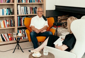 A man speaks with a woman in the hotel library while a microphone is set up for the podcast recording.
