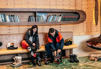 Two guests are putting on ski boots in a cosy wooden lounge, with a helmet, gloves and boots beside the bench.