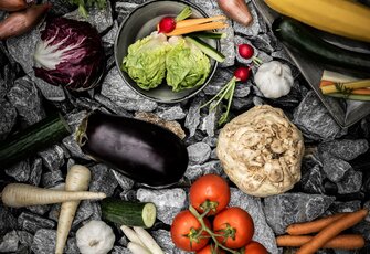 Verschiedene Gemüse wie Tomaten, Auberginen, Radicchio, Sellerie und Karotten liegen auf dunklen Steinen.