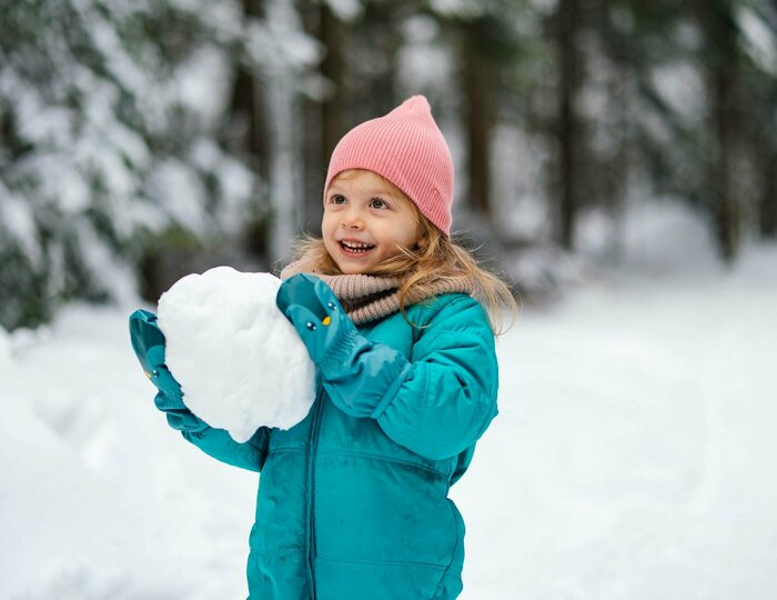 Ein Kind im türkisfarbenen Wintermantel hält draußen im Schnee einen Schneeball in der Hand.