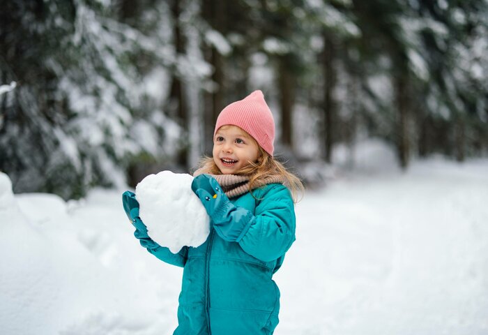 Ein Kind im türkisfarbenen Wintermantel hält draußen im Schnee einen Schneeball in der Hand.