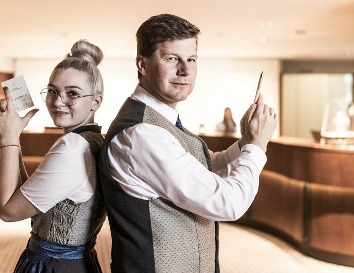 Zwei Mitarbeitende stehen in der Hotelrezeption Rücken an Rücken und halten ihre Arbeitsutensilien in der Hand.