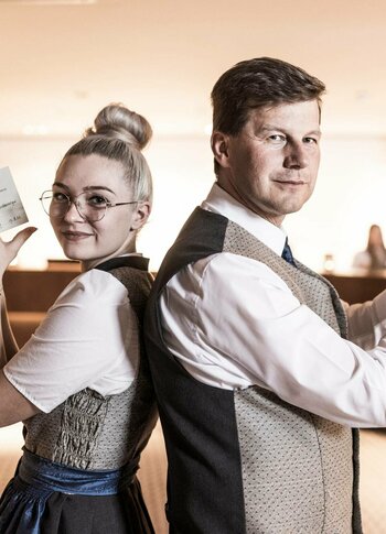 Rezeptionsteam im Hotel Hochschober Zwei Mitarbeitende stehen in der Hotelrezeption Rücken an Rücken und halten ihre Arbeitsutensilien in der Hand.
