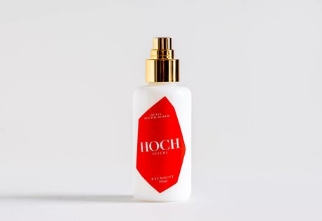 A white spray bottle with a gold cap and red label stands on a light background.