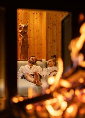Ein Paar entspannt in einer gemütlichen Sauna-Lounge, während im Vordergrund ein Feuer flackert.