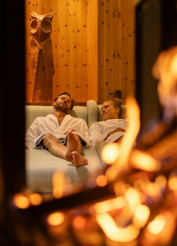 Ein Paar entspannt in einer gemütlichen Sauna-Lounge, während im Vordergrund ein Feuer flackert.