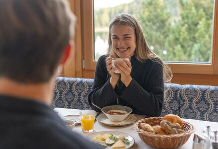 Eine Frau lächelt beim Frühstück im Hotelrestaurant, vor ihr stehen Müsli, Orangensaft und ein Brotkorb.