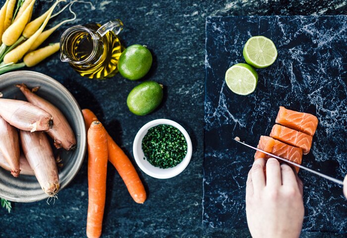 Auf einer dunklen Arbeitsfläche liegen Lachs, Schalotten, Karotten, Limetten, Öl und Kräuter für die Zubereitung bereit.