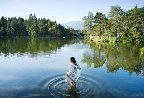 Eine Frau steht im flachen Wasser eines Sees, umgeben von Wald und Bergen.