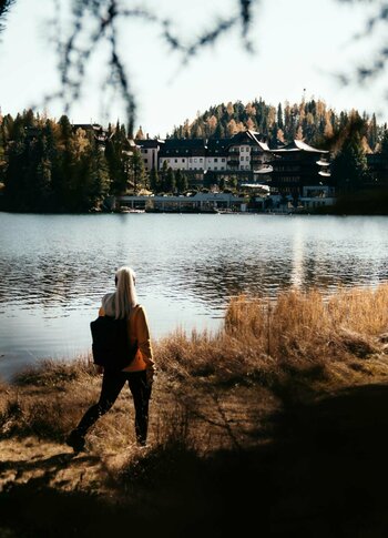Eine Person wandert am Ufer des Sees, gegenüber liegt das Hotel Hochschober zwischen herbstlichen Bäumen.