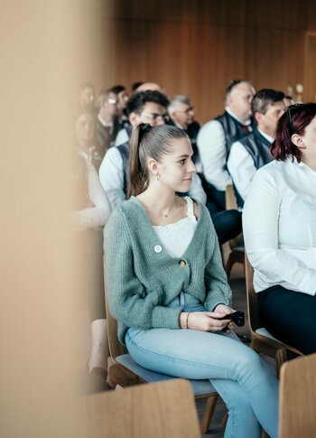 Eine junge Frau sitzt in einer Seminarveranstaltung auf einem Stuhl, im Hintergrund hören weitere Teilnehmende zu.