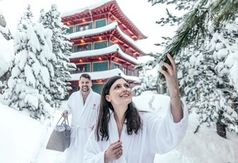 Eine Frau und ein Mann im Bademantel stehen im Schnee vor dem Chinaturm des Hotel Hochschober.