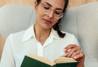 Eine Frau liest entspannt in einem Lesesessel mit hellem Bezug und weißer Bluse.