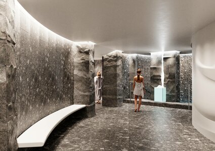 A modern spa interior featuring smooth stone walls and a curved layout. Two women in bathrobes are present, one walking towards a light blue feature, while the other stands nearby. Soft lighting enhances the serene atmosphere, and a white bench is visible along the wall.