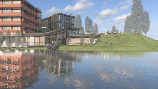 A modern building complex featuring a mix of glass and brick architecture, situated by a calm lake. Lush green landscaping with trees surrounds the area, and a gentle slope leads down to the water's edge. The sky is partly cloudy, creating a serene atmosphere.