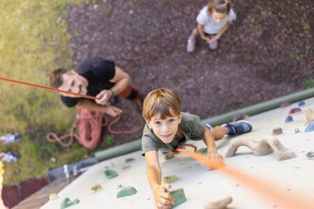 Ein Junge klettert an einer Kletterwand, während zwei weitere Kinder und ein Erwachsener von unten zusehen.