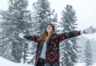 Eine Frau steht im Schnee vor verschneiten Bäumen und streckt die Arme aus.