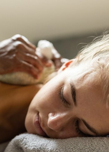 A woman lies with closed eyes on a treatment table while receiving a wellness massage.