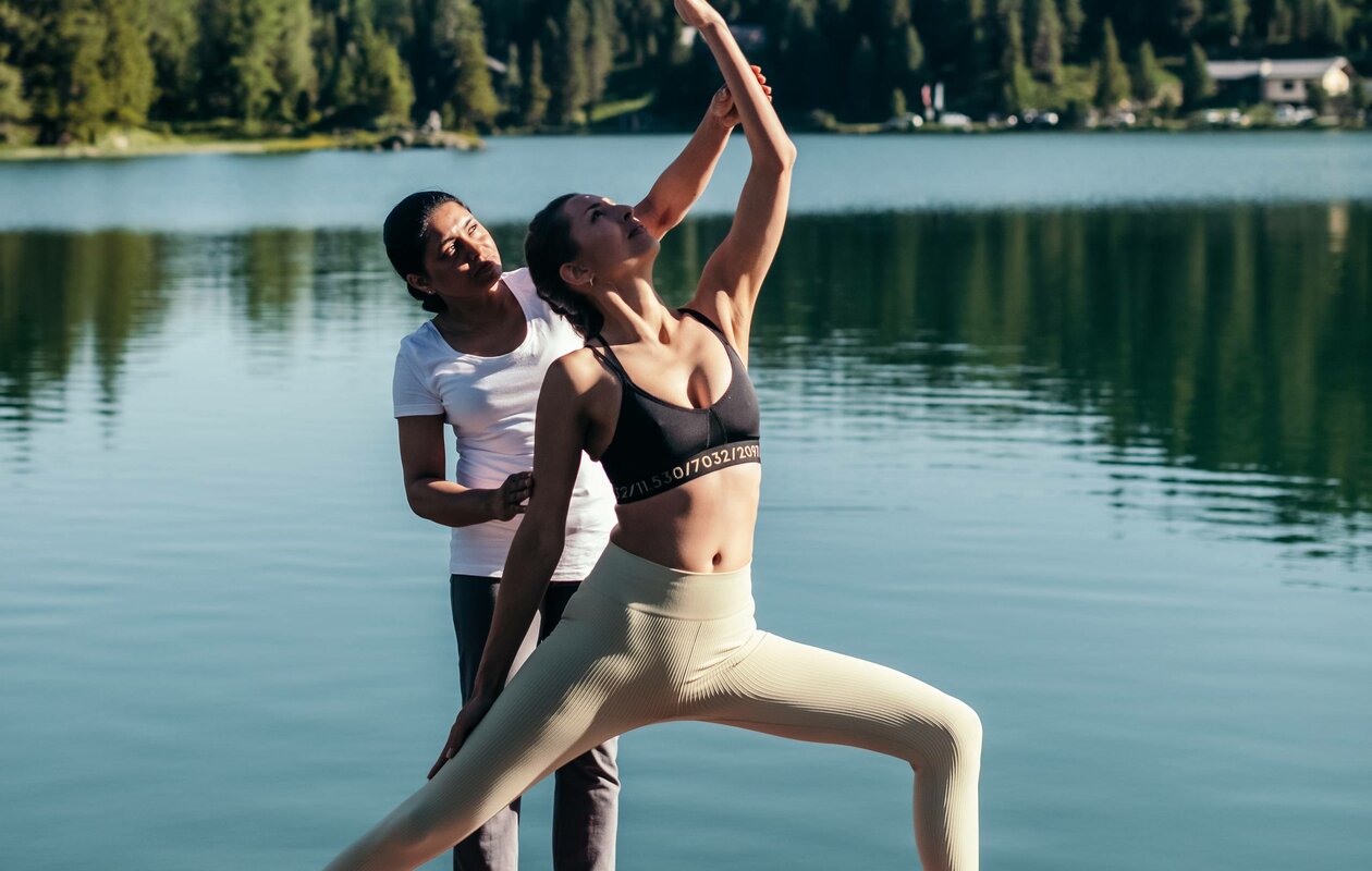 Eine Frau übt Yoga am Seeufer, während eine zweite Person sie im Hotel Hochschober unterstützt.