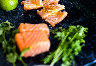 Mehrere Stücke frisches Lachsfilet liegen auf einer dunklen Platte mit grünem Gemüse und einer Limette.