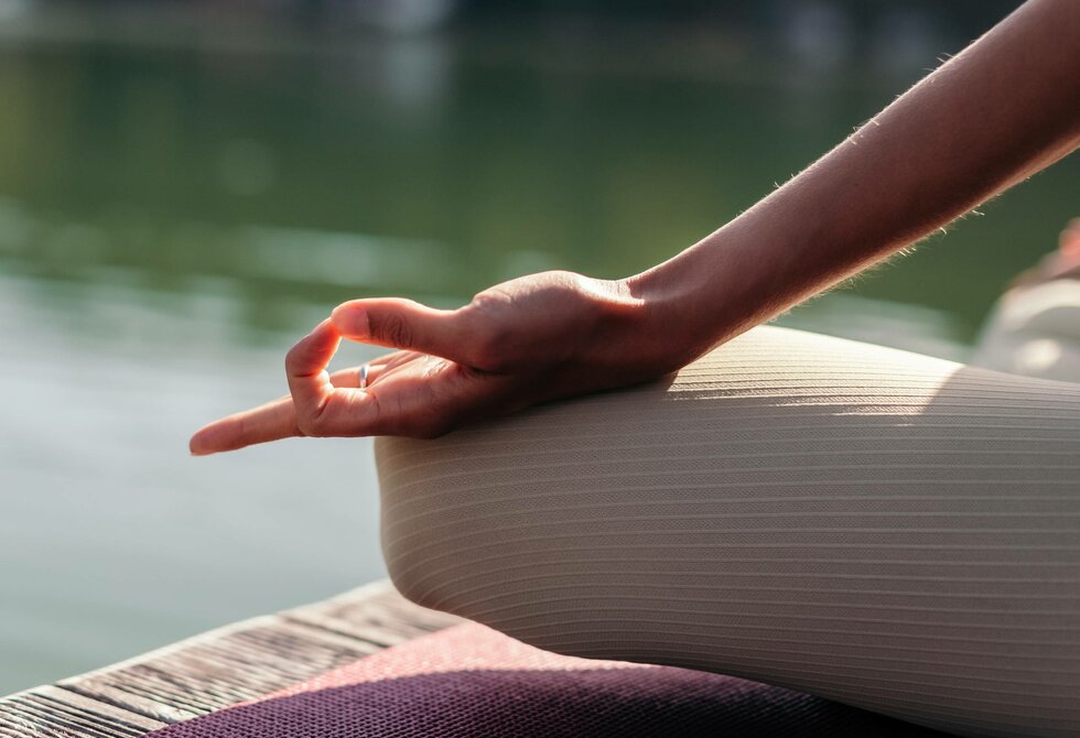 Eine Person sitzt im Yoga-Sitz am Seeufer und hält die Hand in einer Meditationsgeste auf dem Knie.