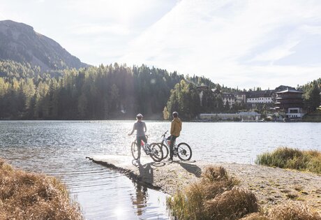 Zwei Gäste stehen mit ihren Fahrrädern auf einem Steg am Seeufer vor bewaldeten Bergen.