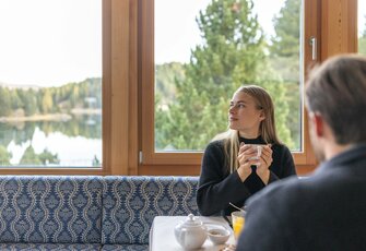 Eine Frau sitzt mit einer Tasse am Tisch im Hotelrestaurant, während im Hintergrund der See und Bäume zu sehen sind.