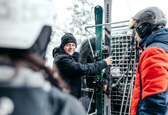 A man in ski gear secures his skis at a lift station while two other people stand in the foreground and on the right.