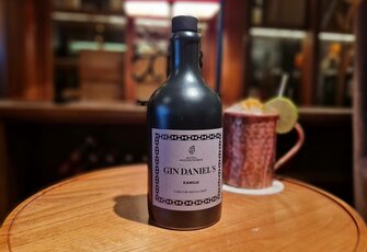 A black gin bottle stands on a wooden table beside a copper mug with a drink and a lemon slice.