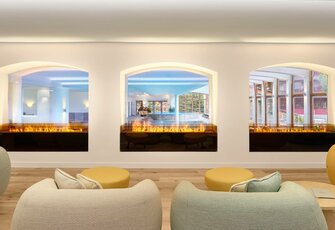 Eine helle Lounge mit Polstermöbeln, runden Tischen und drei großen Fensteröffnungen mit Feuerspiel davor.