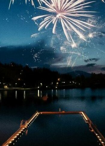 Feuerwerk über dem See bei Nacht mit beleuchteter Badeplattform im Vordergrund.