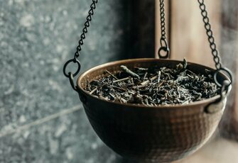 A hanging metal bowl filled with herbs stands by the sauna wall and the warm wooden window frame.