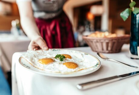 Eine Person serviert ein Frühstücksgericht mit Eiern auf einem weiß gedeckten Tisch.
