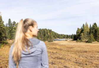 Eine Frau steht unscharf im Vordergrund und blickt über eine herbstliche Wiese zum Turracher See zwischen den Nockbergen.