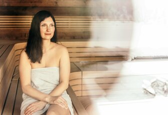 Eine Frau sitzt in der Sauna auf einer Holzbank und blickt ruhig zur Seite.