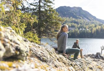Zwei Personen sitzen auf einem Felsen am Ufer des Turracher Sees vor einer Berglandschaft.