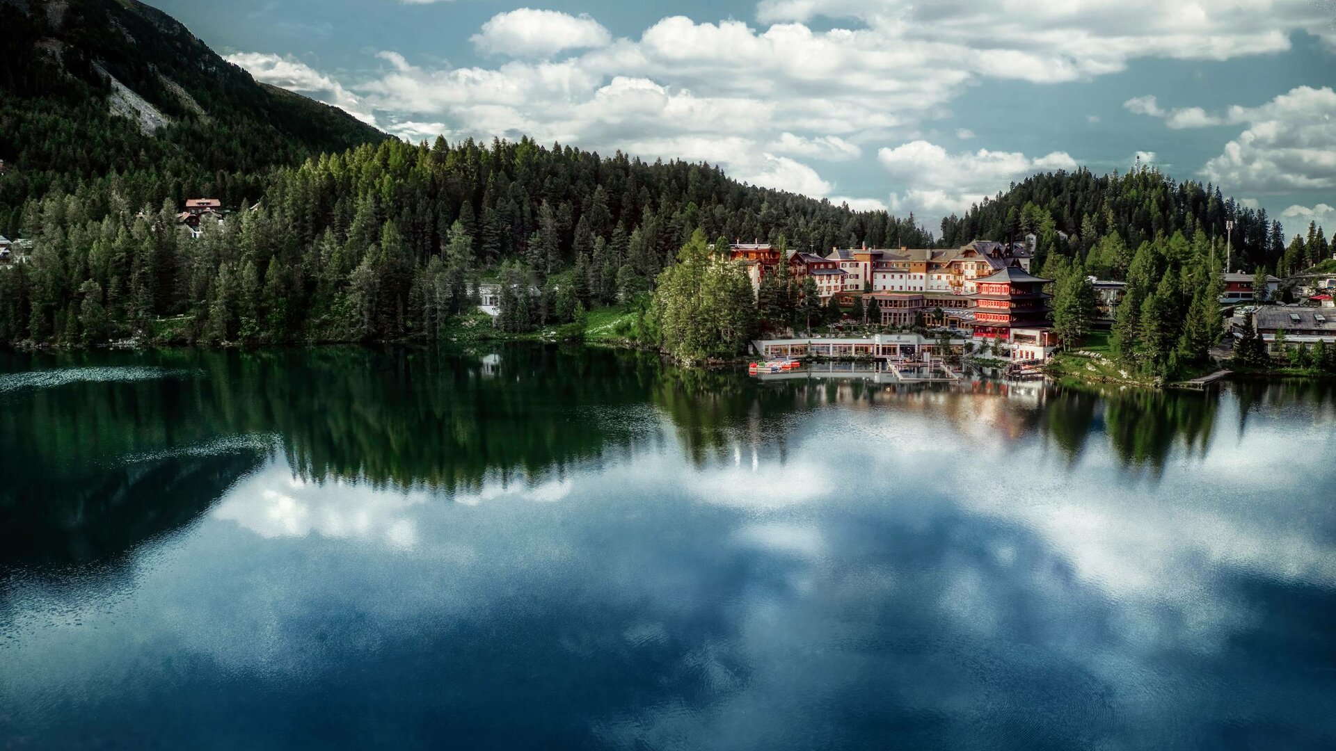Lake Turracher See reflects the forest and Hotel Hochschober on the shore beneath a cloudy sky.