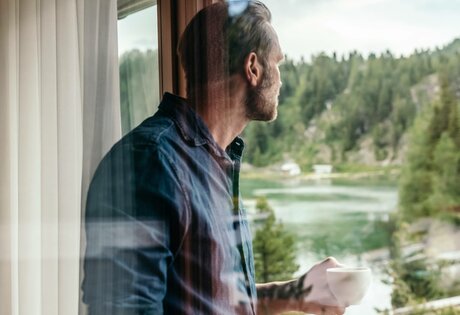 Ein Mann steht am Fenster und blickt mit einer Tasse in der Hand auf den See und den Wald draußen.