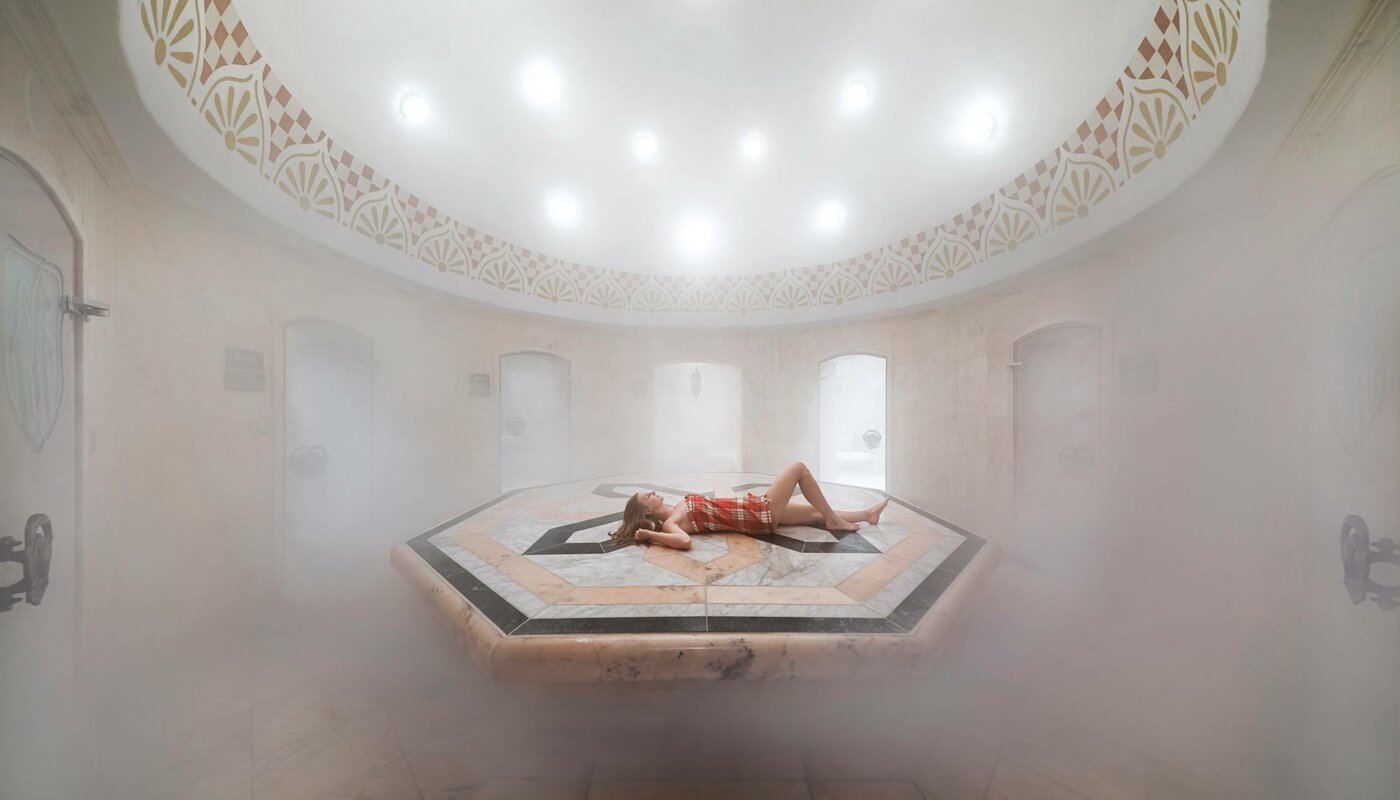 A person lies relaxed on the raised stone platform in the oriental hamam, surrounded by dense steam.