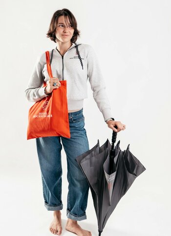 A woman holds an orange Hochschober bag and a black umbrella against a light background.